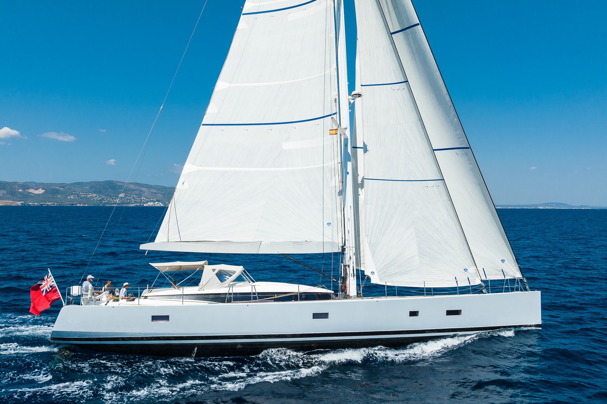 Modern sailing yacht with full white sails cruising near coast under clear blue sky