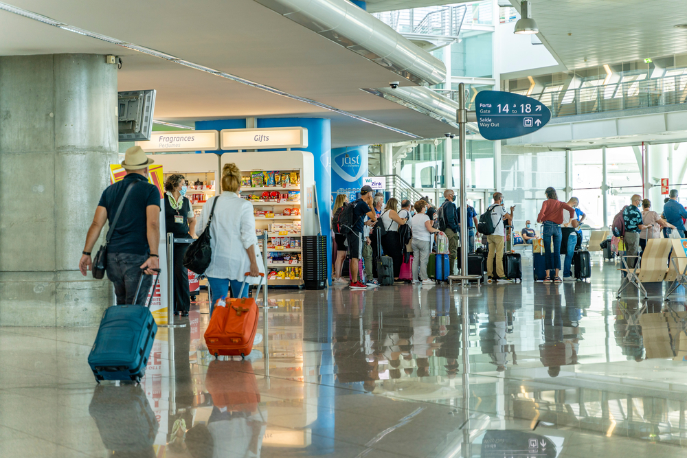 Travellers with luggage walk through Lisbon Airport past shops and gate signs
