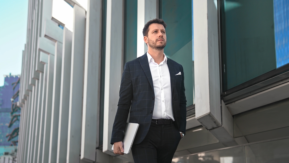 Confident, serious business man in suit walking city street holding laptop