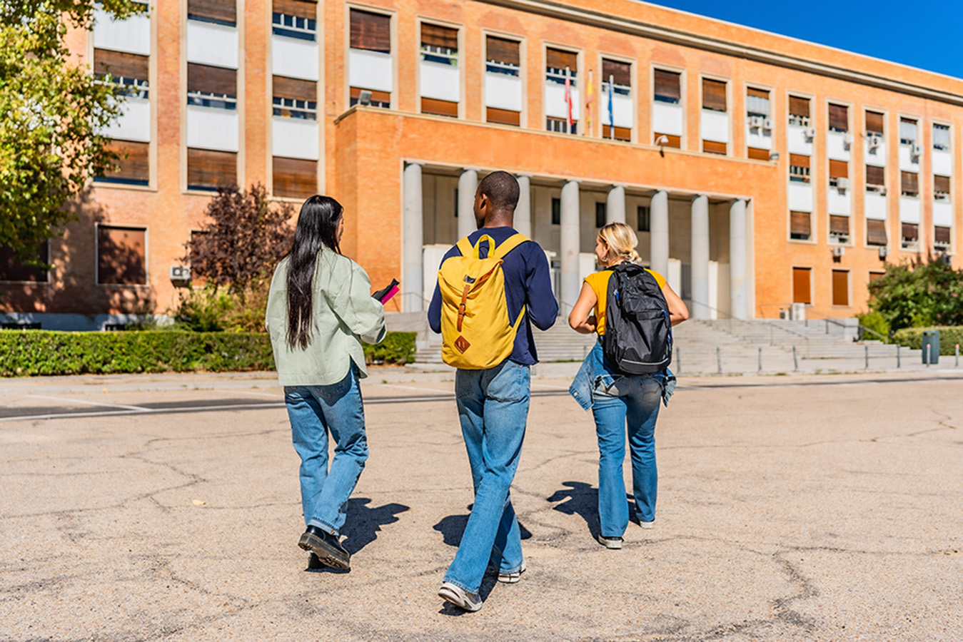 Three students with backpacks walk toward a large brick university building in the sun