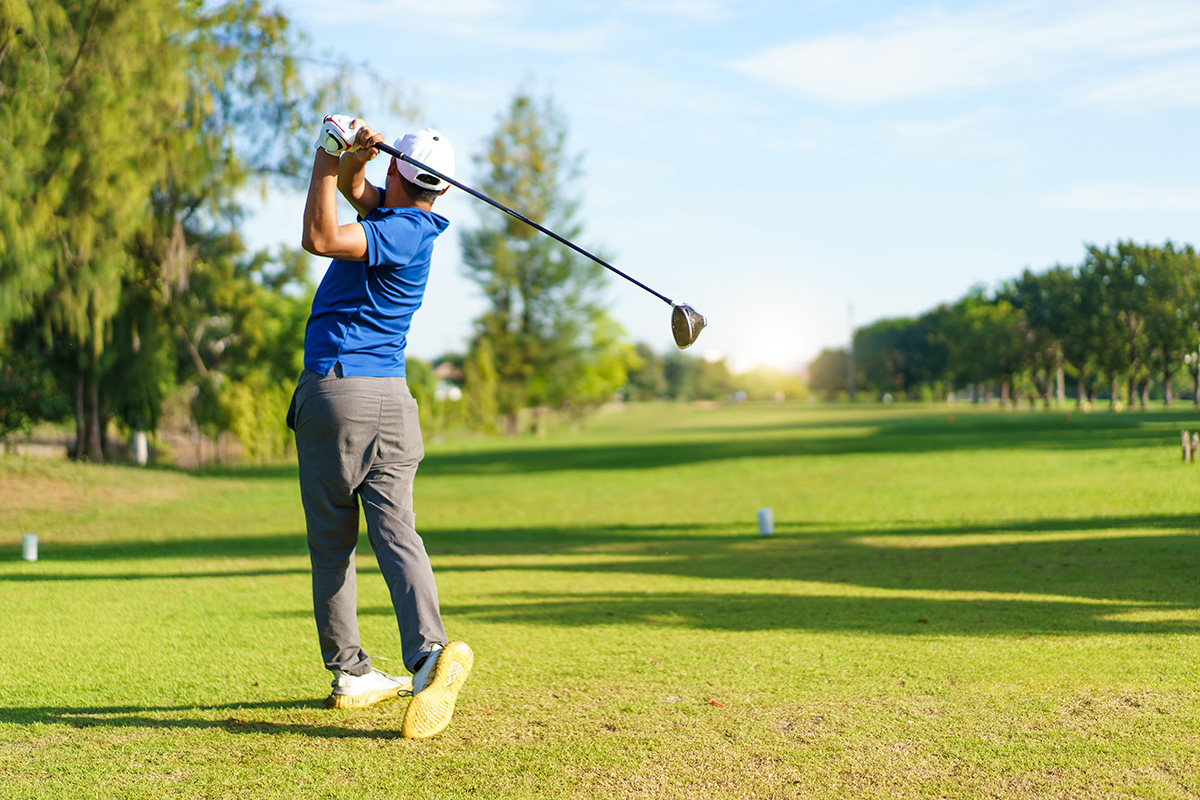 A golfer in a blue shirt completes a swing with a driver on a sunny, green golf course