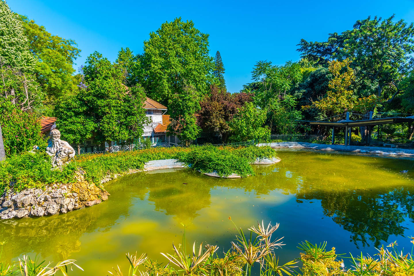 Scenic park pond in Lisbon featuring a stone statue, lush green trees, and clear blue sky