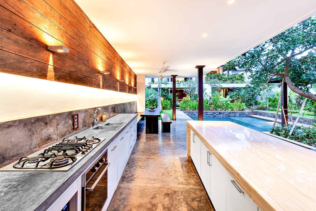An outdoor kitchen with a long concrete counter, wooden wall, and view of a pool