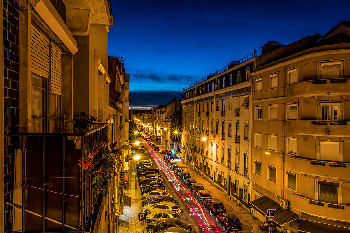 A long exposure night view of a Lisbon street with glowing car light trails and warm lamps