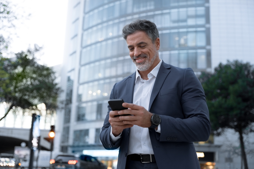 Male investor conducting business from mobile phone outside of modern high-rise building