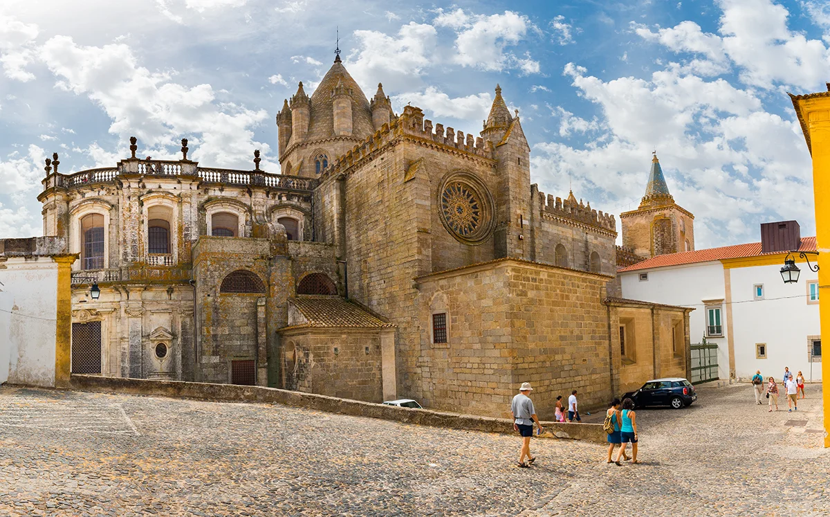 Tourists stroll past the historic stone facade and towers of the Cathedral of Évora