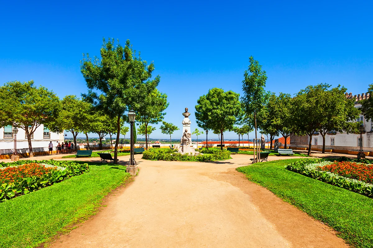 A lush green park with manicured lawns, leafy trees, and a central stone monument statue