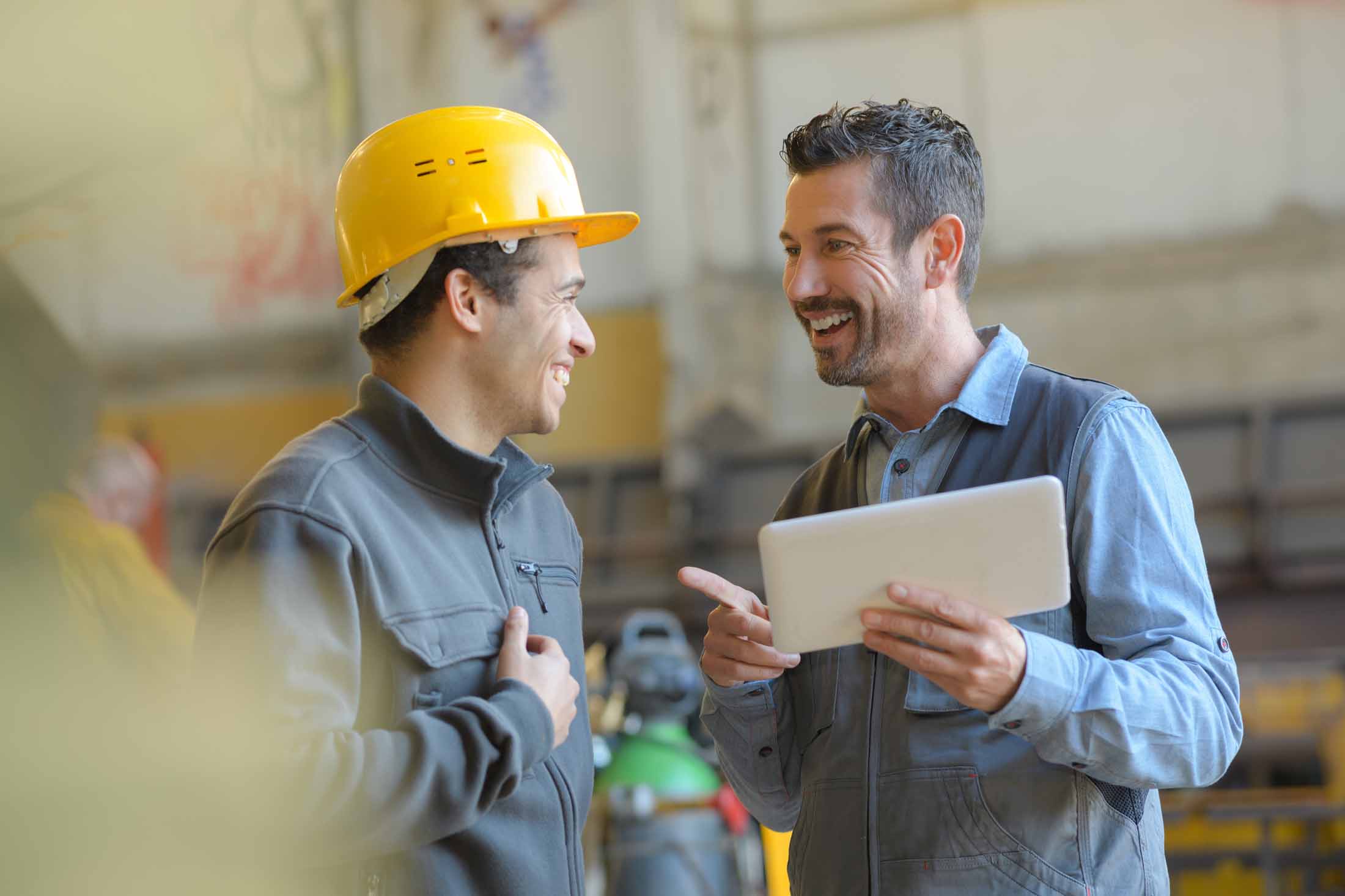 Two men in a workshop smiling and talking, one wearing a yellow hard hat and the other holding a tablet.
