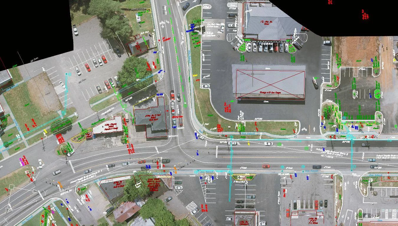 Aerial view of a busy intersection with multiple lanes, traffic signals, parking lots, and buildings with annotations and markings in various colors.