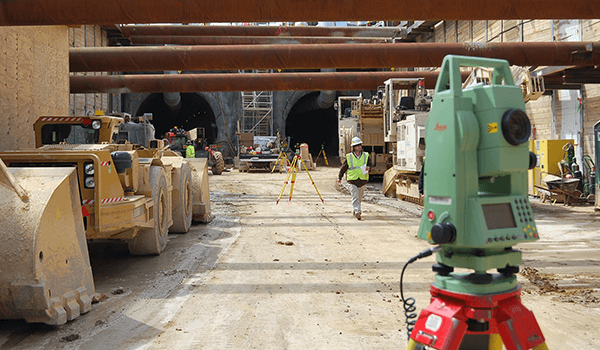Construction site inside a tunnel with heavy machinery, surveying equipment, and a worker wearing a safety vest and helmet.