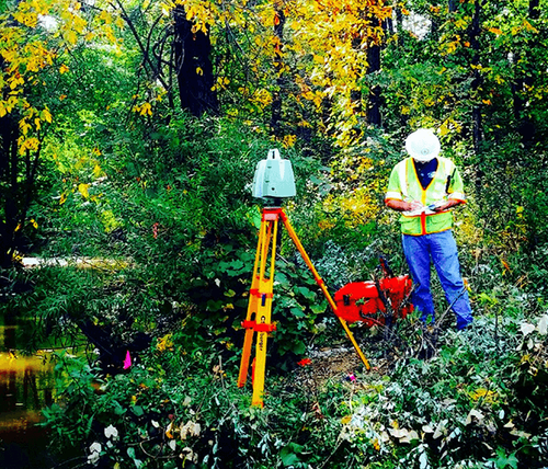 Surveyor wearing a hard hat and safety vest working with surveying equipment in a green and yellow forested wetland area.