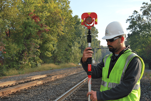 Construction worker in a white hard hat and green safety vest holding a surveying rod near railroad tracks surrounded by trees.