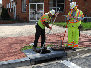 Two construction workers with hard hats and safety vests inspecting and working near an open manhole on a sidewalk.