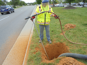Worker wearing high-visibility vest and face shield using a tool to clean or inspect a hole next to a roadside grassy area.