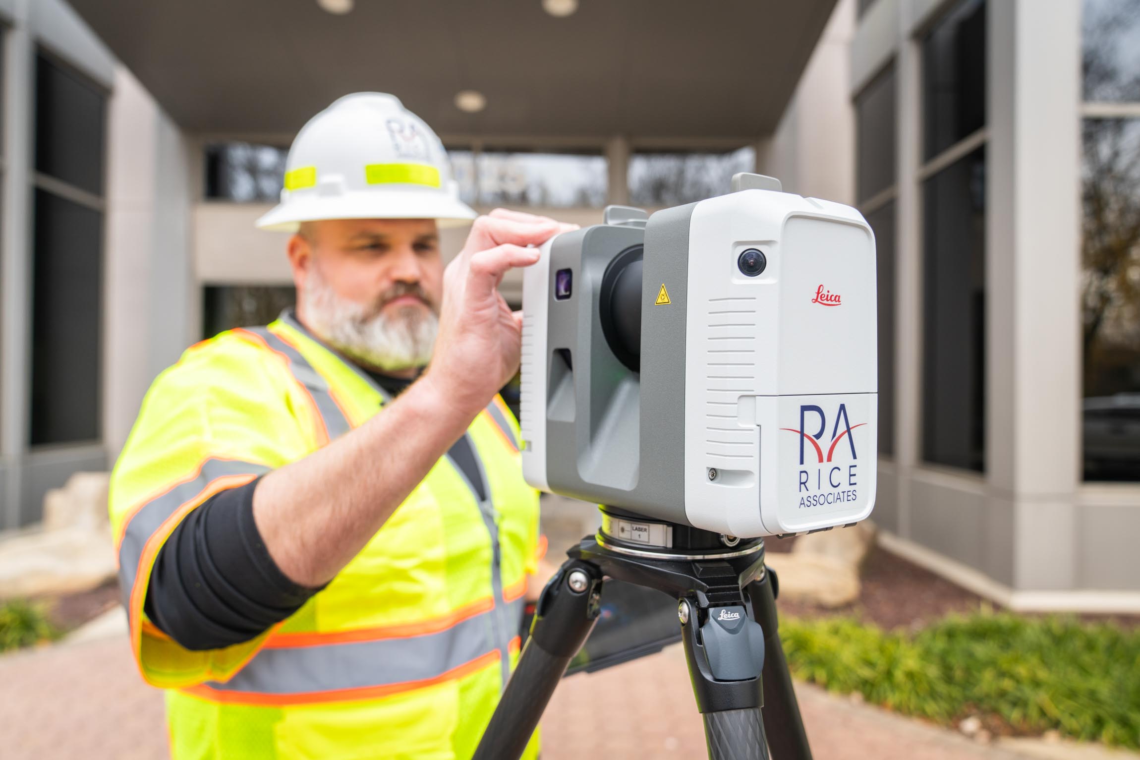 Male construction worker in a hard hat and reflective vest operating a Leica surveying instrument on a tripod outdoors.