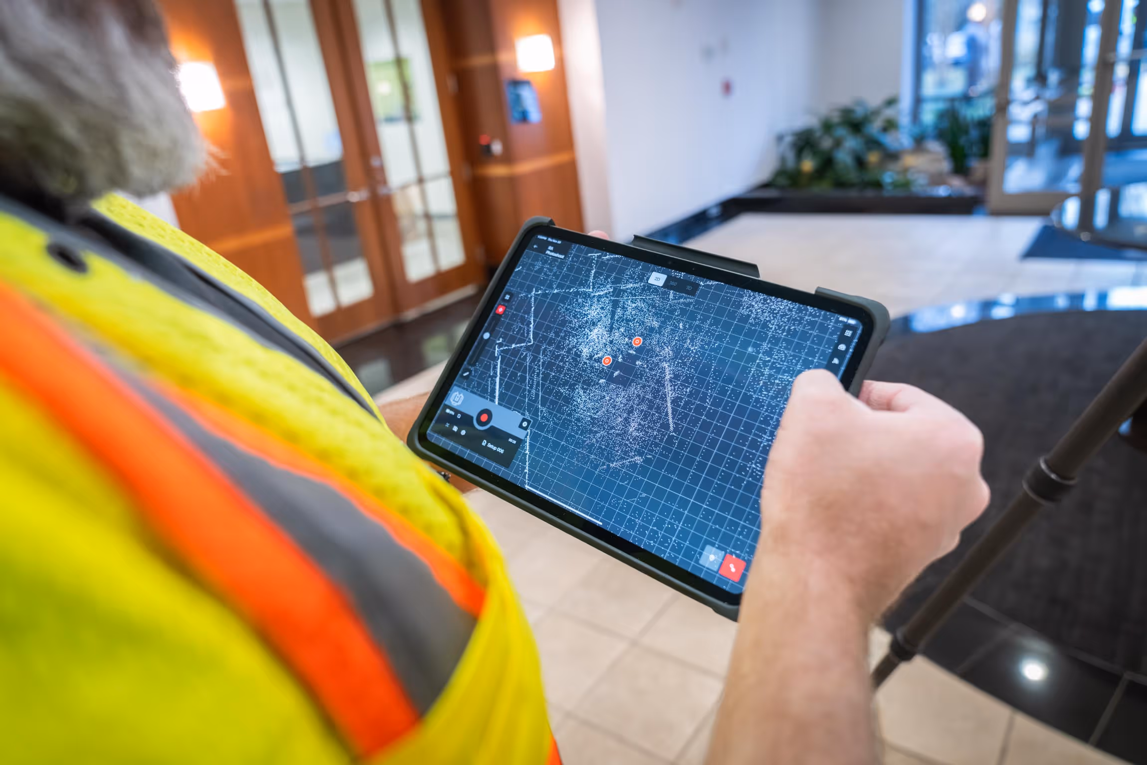 Person in a high-visibility safety vest holding a tablet displaying a digital grid map with scattered white points and markers indoors.