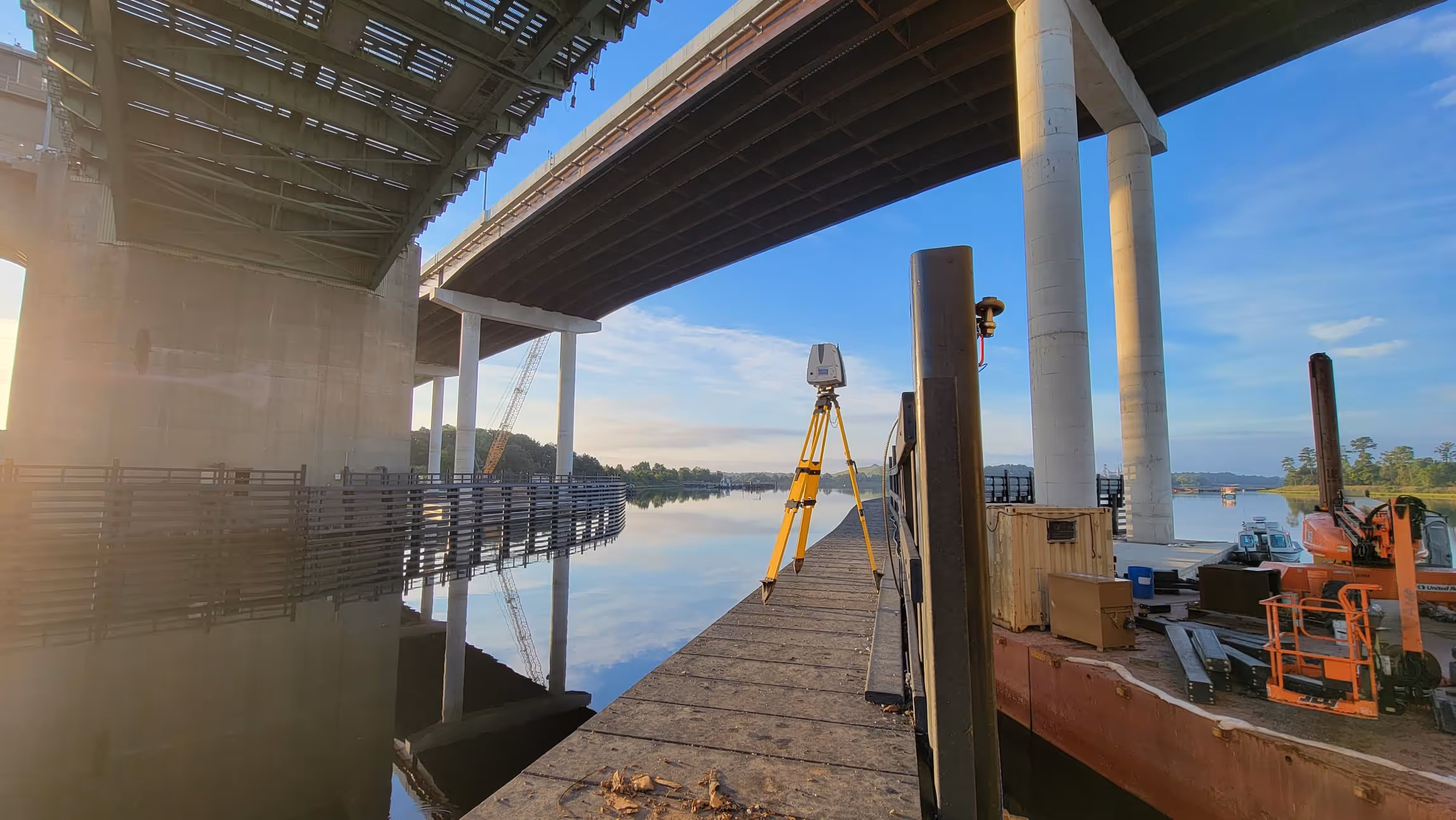 Surveying equipment set up on a dock under a large bridge over calm water reflecting the sky and bridge structures.