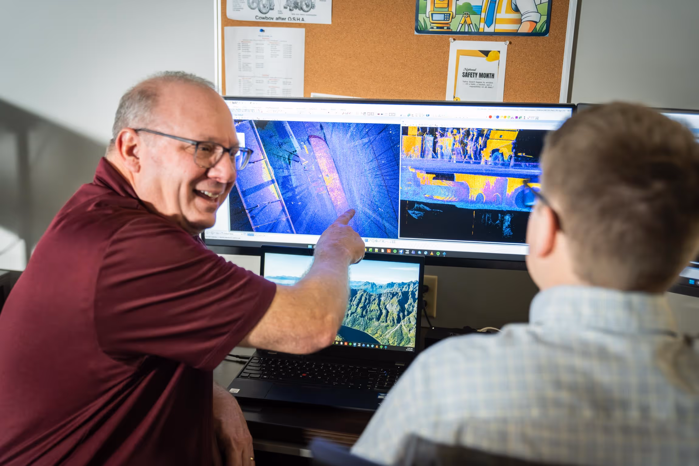 A man in a burgundy shirt pointing at 3D scan images on a computer monitor while talking to another person.
