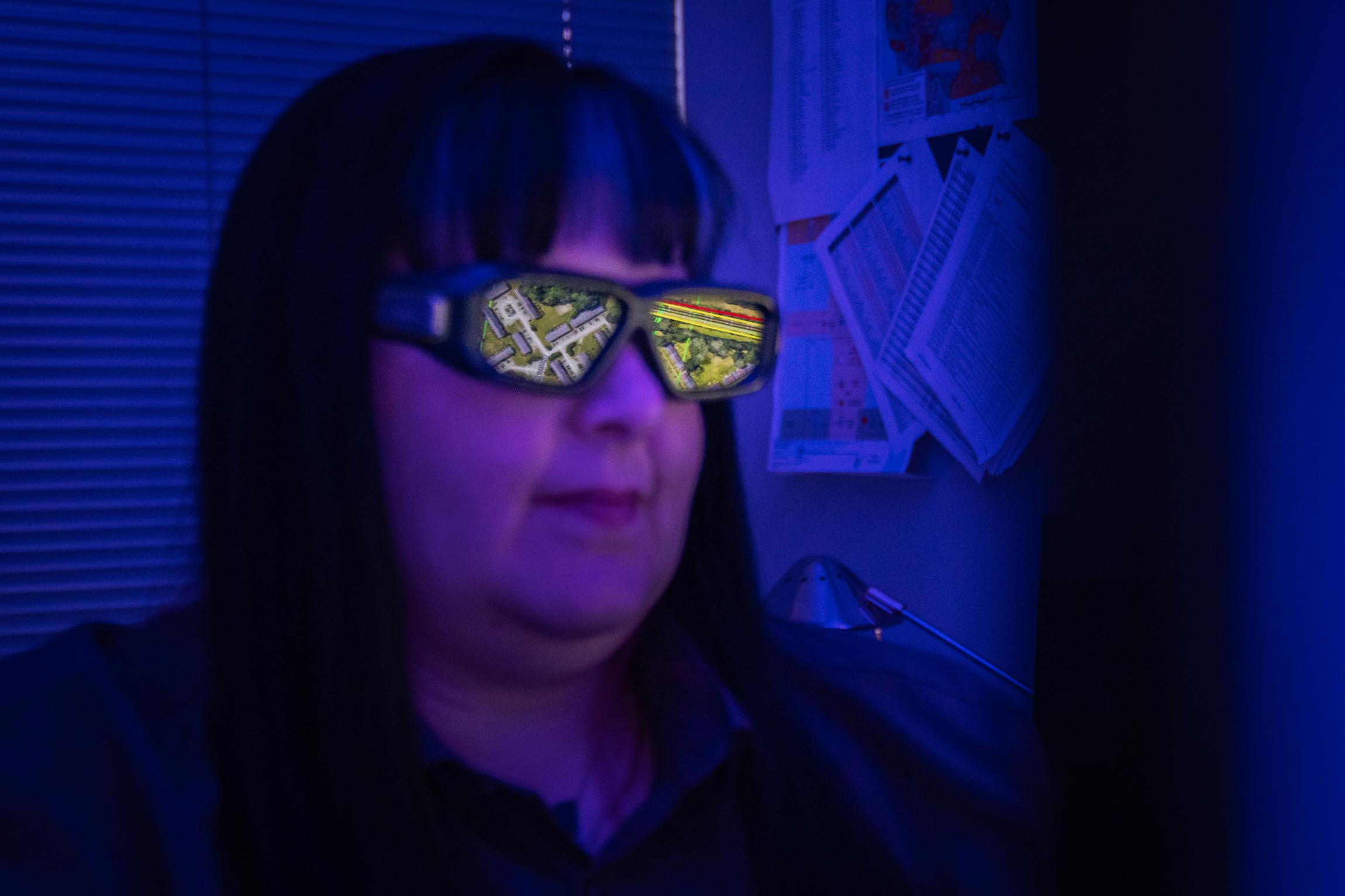 Woman wearing glasses that reflect a detailed aerial map view of roads and buildings, in a dimly lit office.