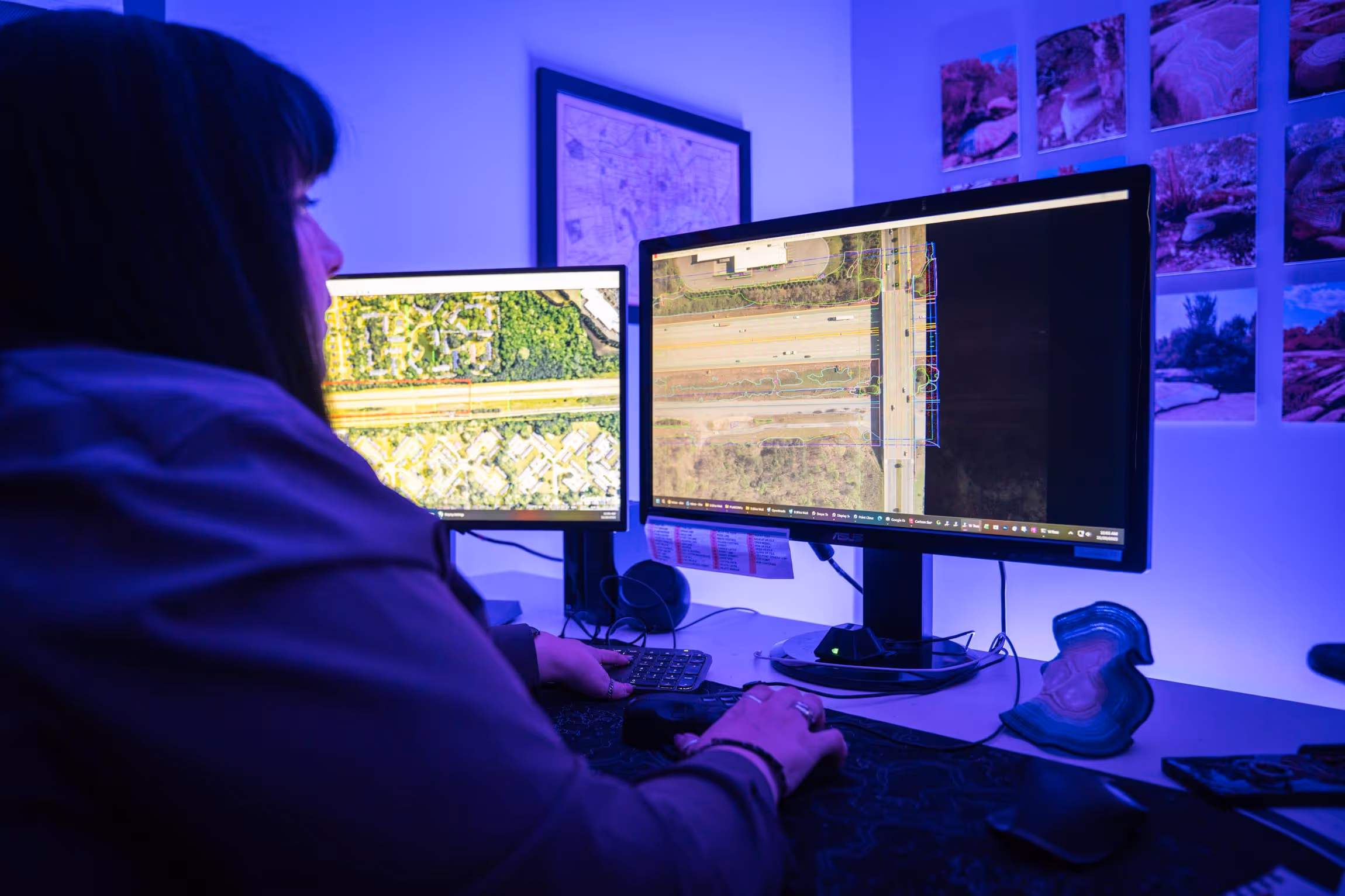 Person working on two computer monitors displaying aerial and map views of roads and surrounding areas in a dimly lit room.