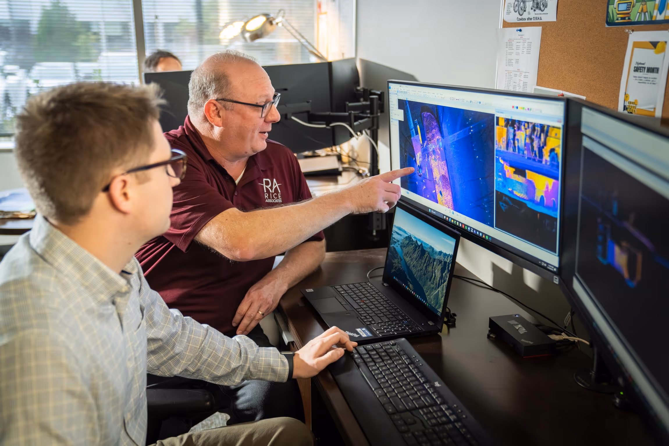 Two men analyze data on a computer with multiple monitors, one pointing to a thermal imaging screen showing colorful heat maps.