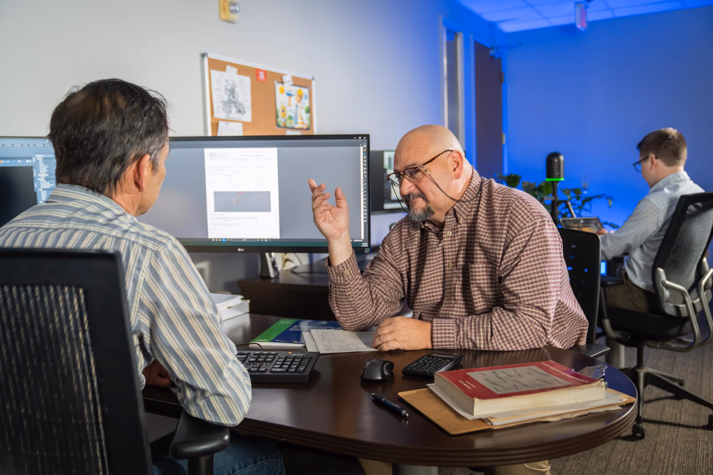 Two men engaged in a discussion at a desk with computer monitors, books, and documents, while another person works on a laptop in the background with blue lighting.
