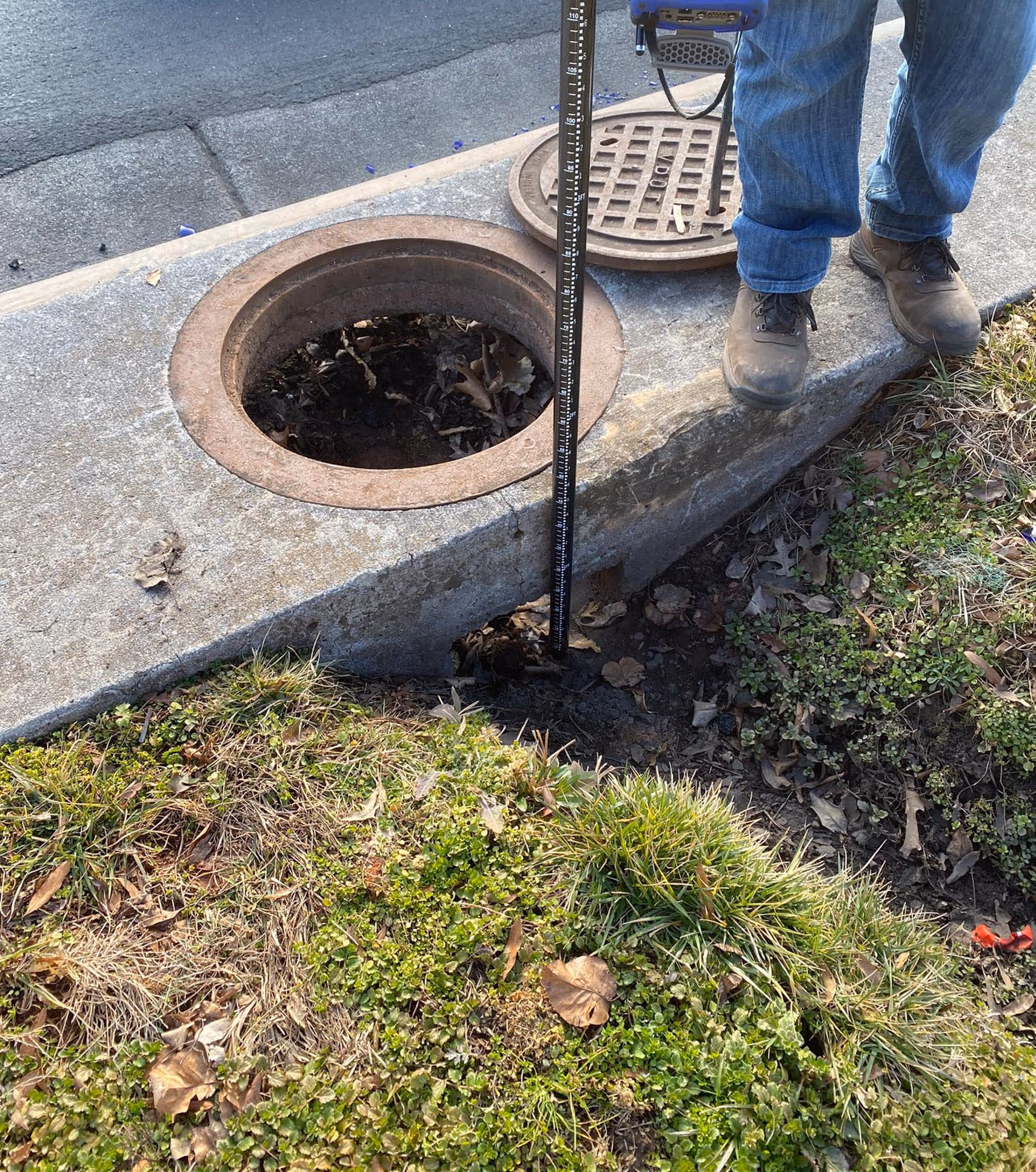 Person standing next to an open manhole with a measuring rod extended into the uncovered hole beside a street curb and grassy area.
