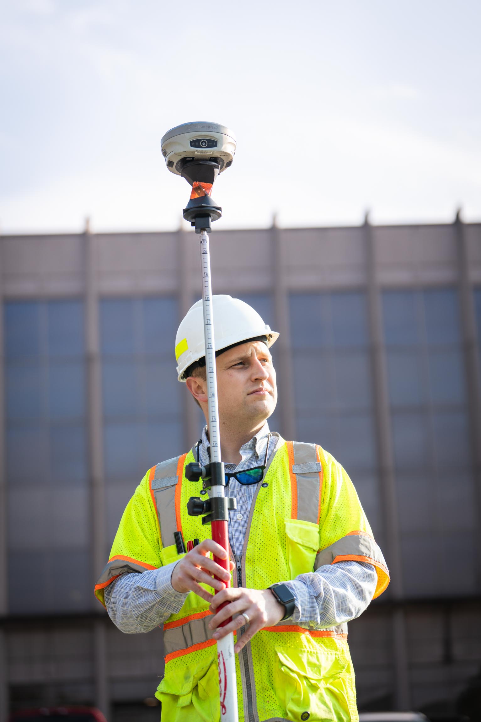 Construction worker in a white hard hat and yellow safety vest holding surveying equipment with a building in the background.