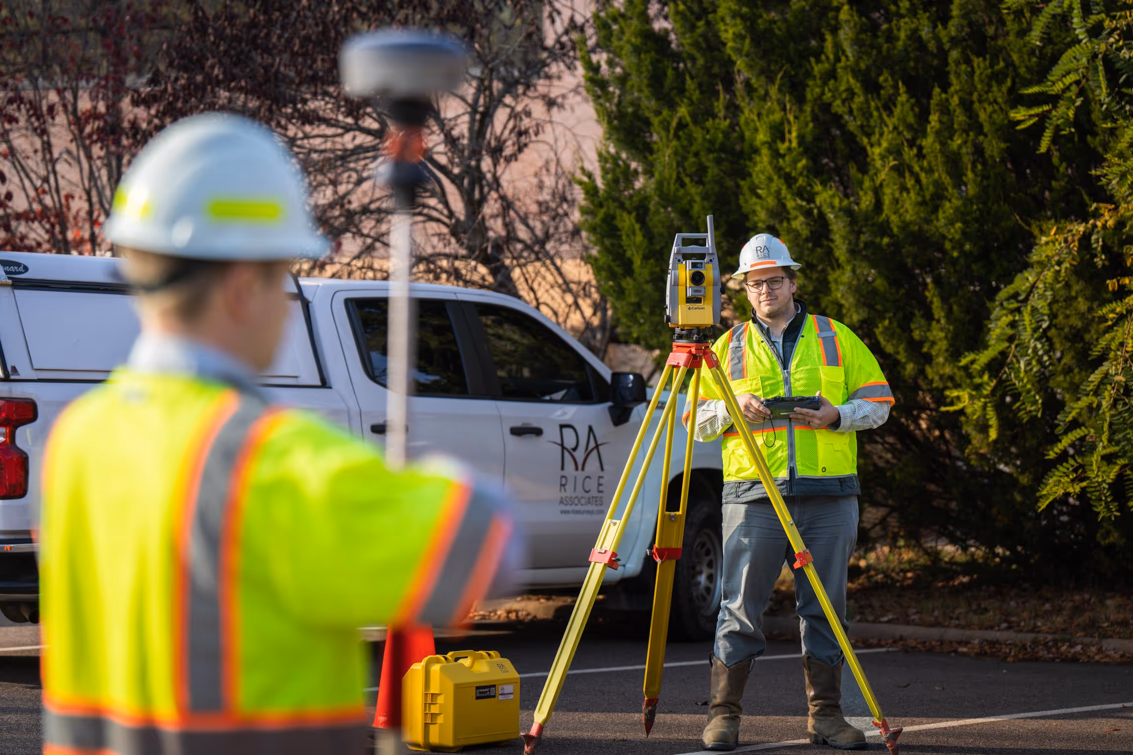 Two surveyors in high-visibility jackets and hard hats using surveying equipment near a white pickup truck with Rice Associates logo.