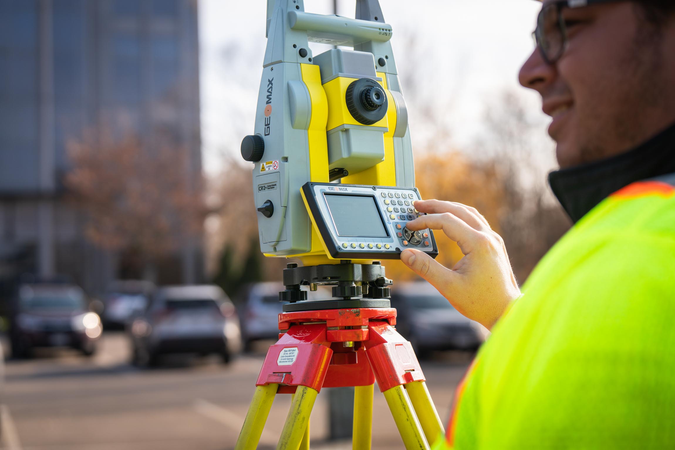 Surveyor in a yellow high-visibility jacket operating a yellow and gray total station instrument on a tripod in an outdoor urban setting.