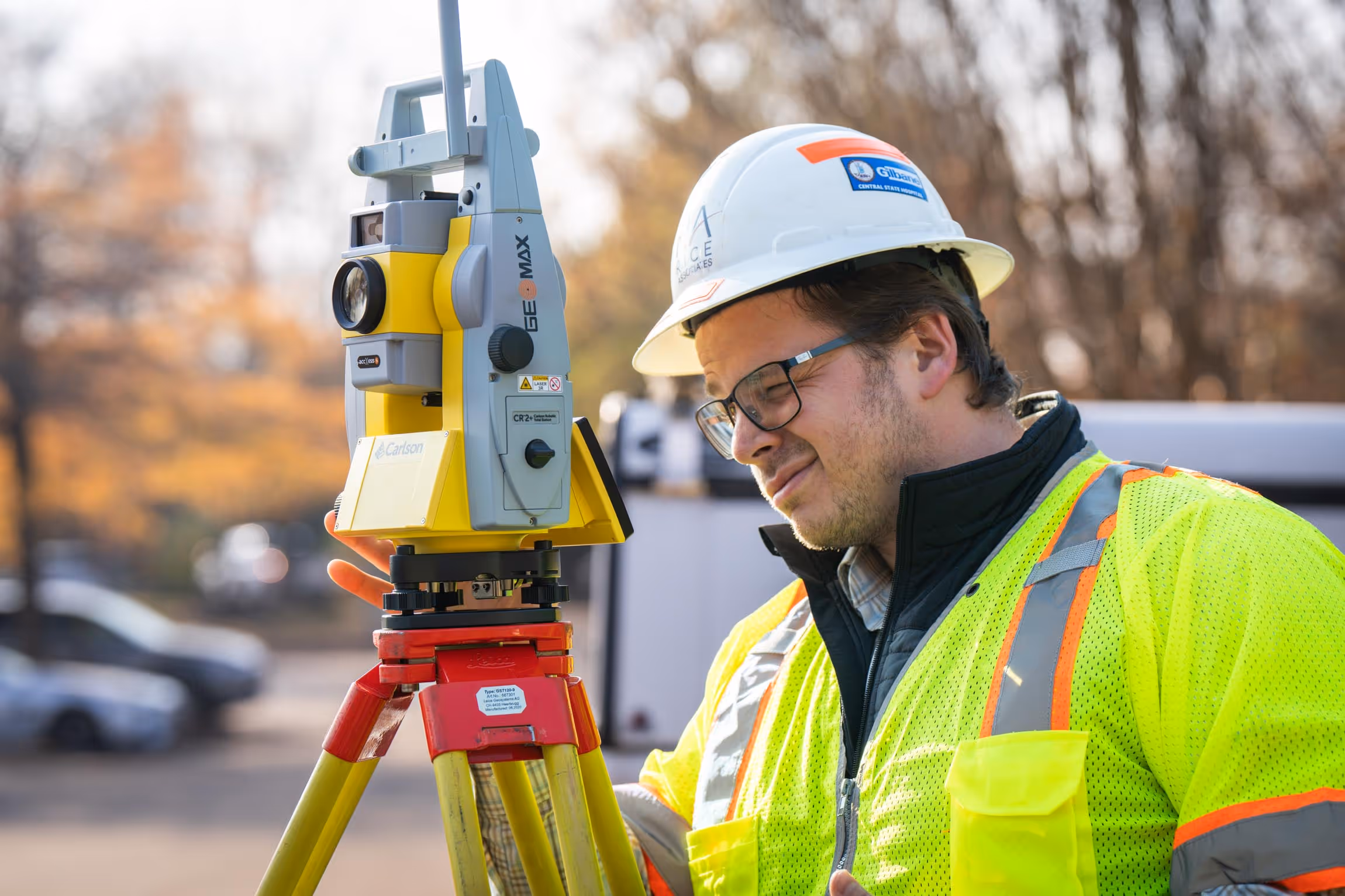 Construction worker in a hard hat and high-visibility jacket using surveying equipment on tripod outdoors.