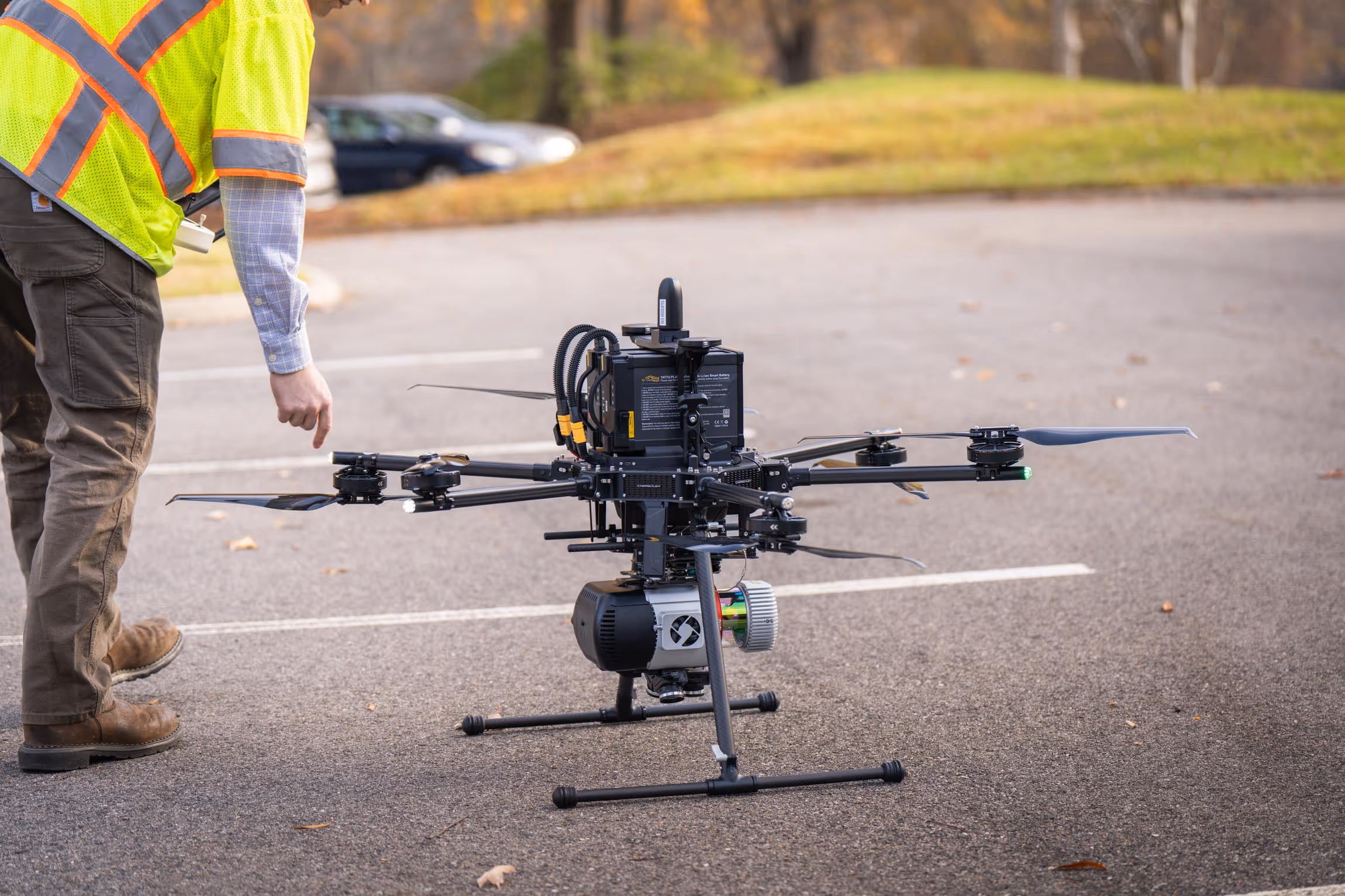 Person in high-visibility vest and brown pants preparing a large black drone with multiple rotors on a paved surface.