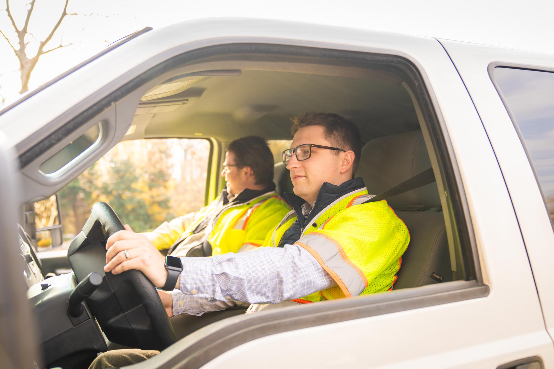 Two men wearing yellow safety vests inside a white vehicle, the man in the passenger seat is looking out, and the driver is focused ahead holding the steering wheel.