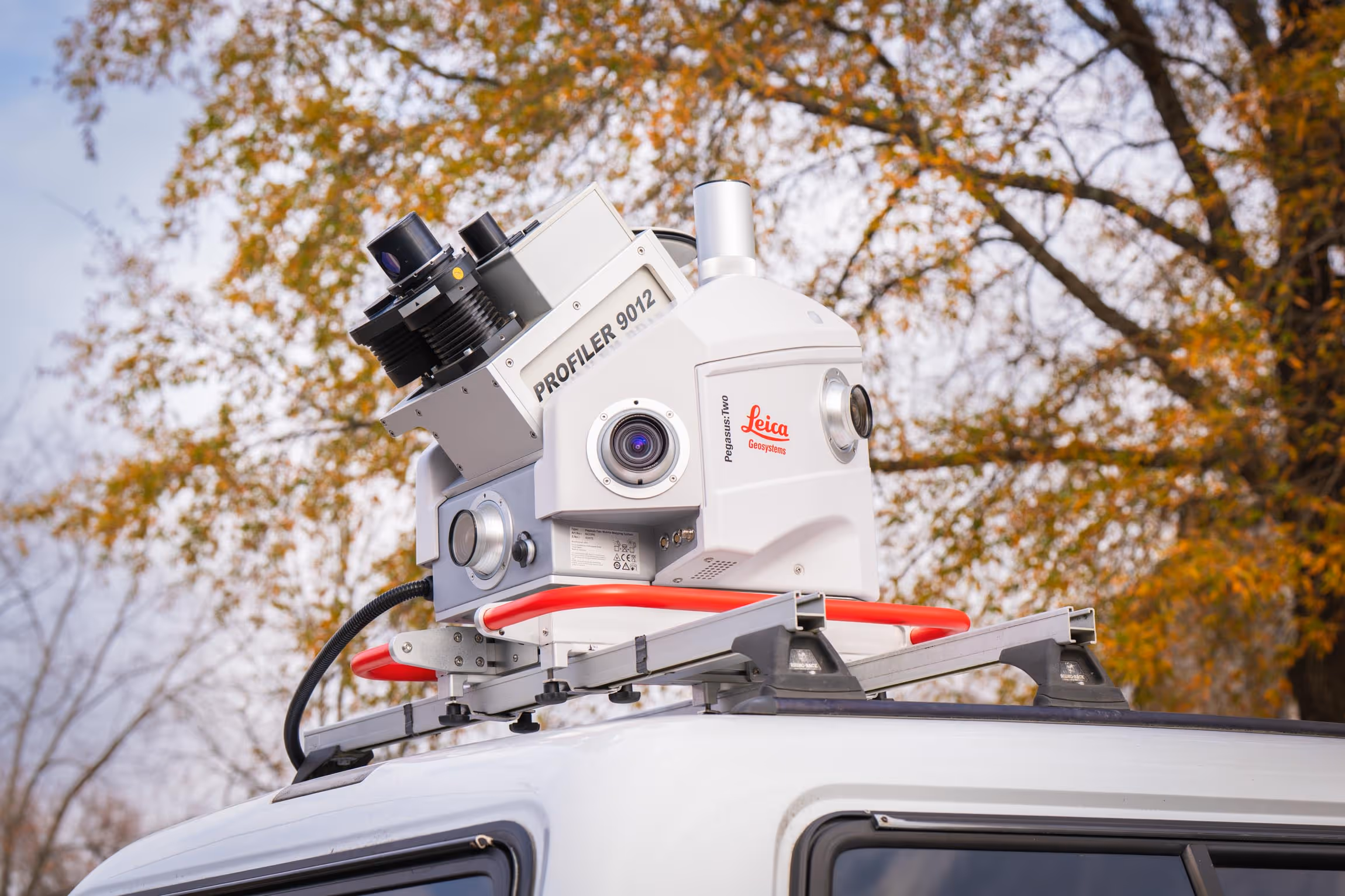 Leica Pegasus Two Profiler 9012 mounted on the roof of a white vehicle against a backdrop of autumn trees.