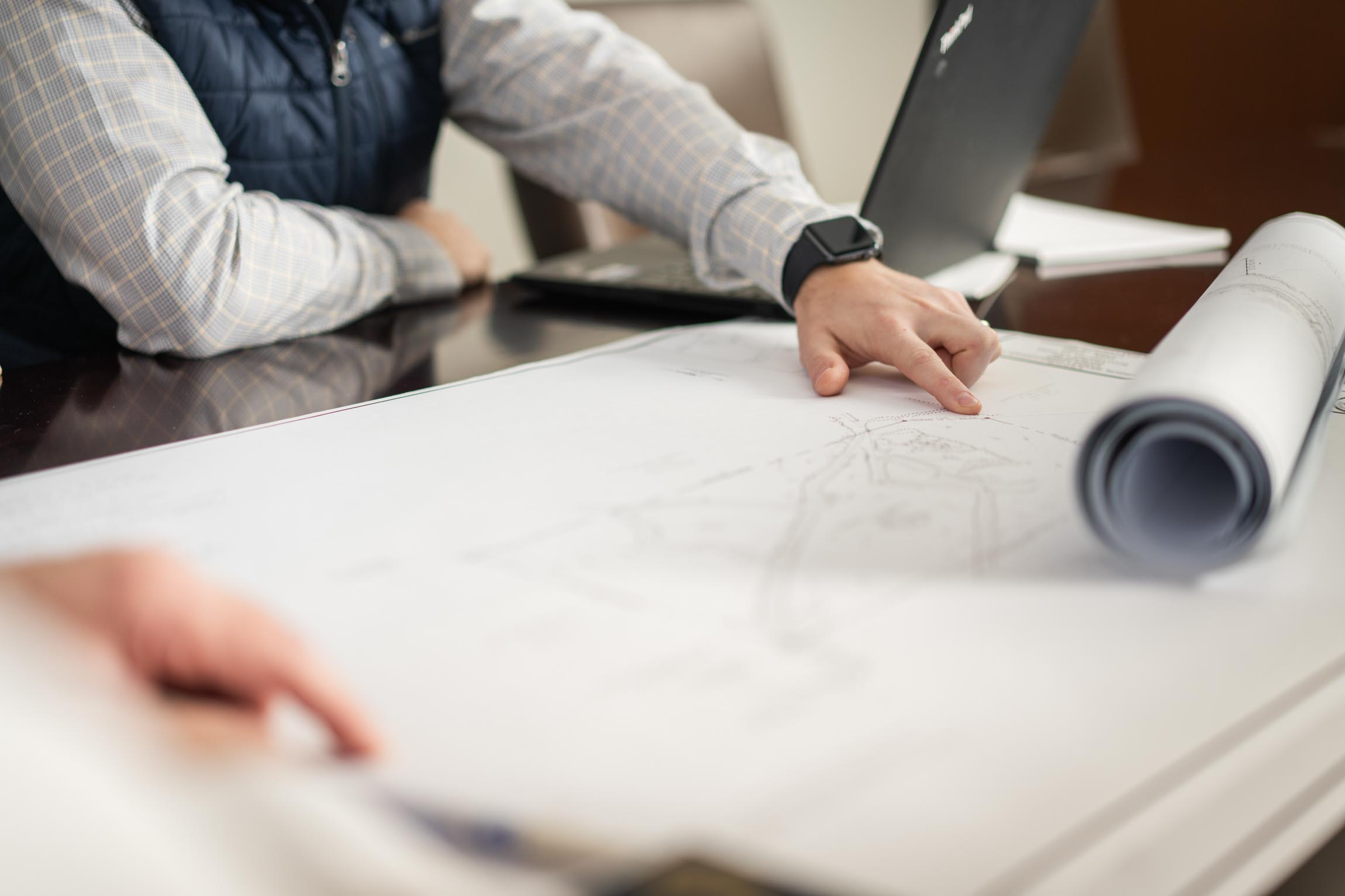 Person pointing at a large blueprint on a table with a laptop in the background.