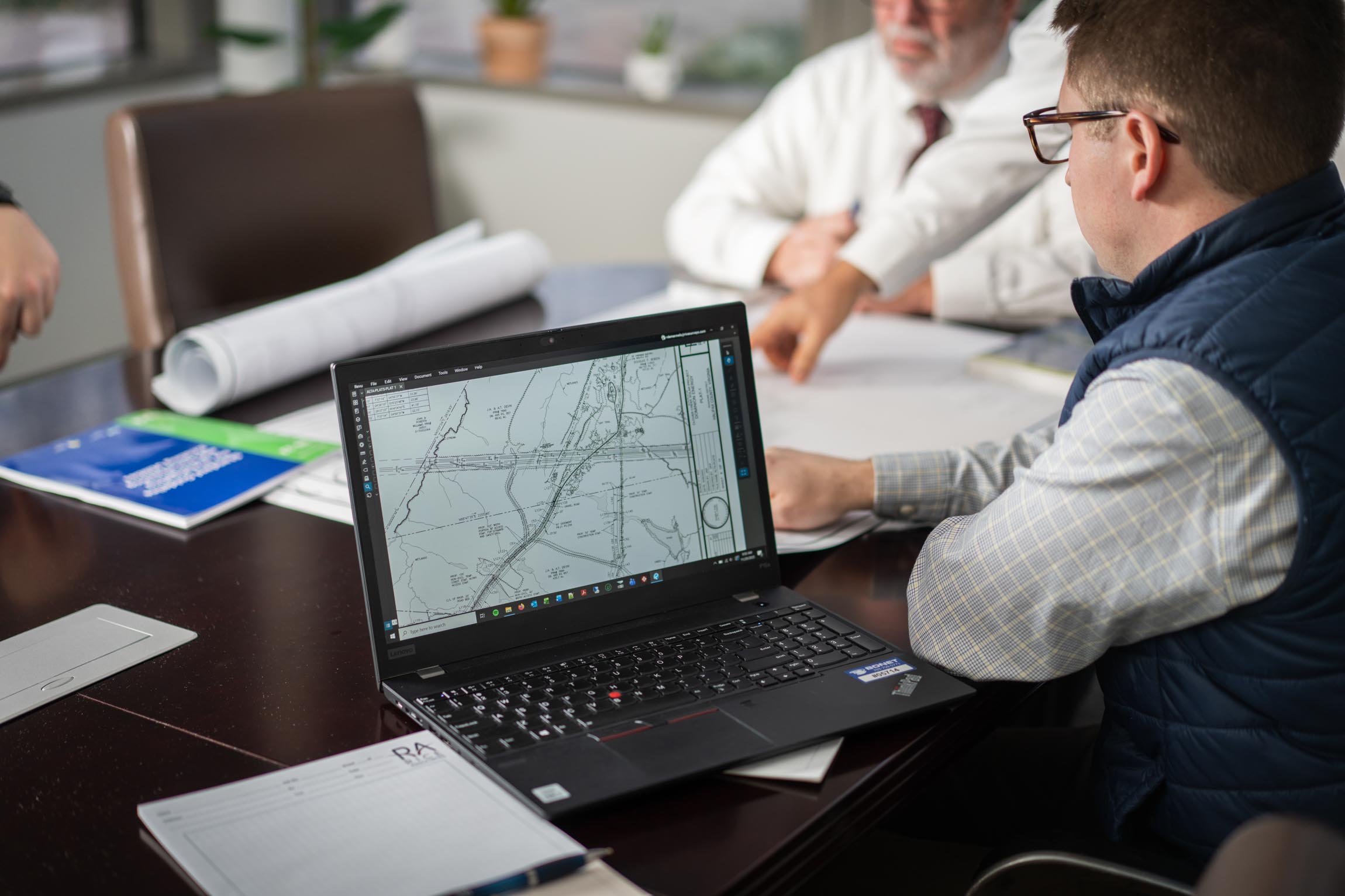 Man in glasses and vest looking at a laptop screen displaying a detailed map in a meeting room with two other men.
