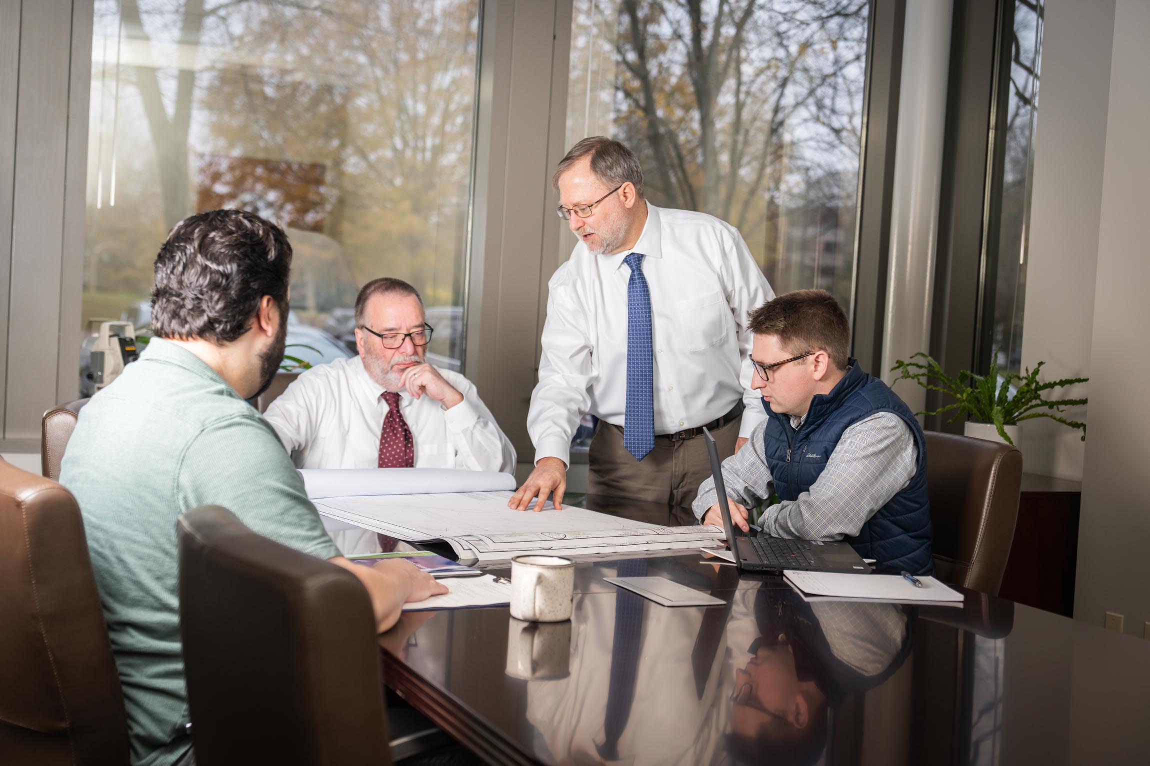 Four men having a business meeting, reviewing large architectural plans on a conference table with a laptop and papers.