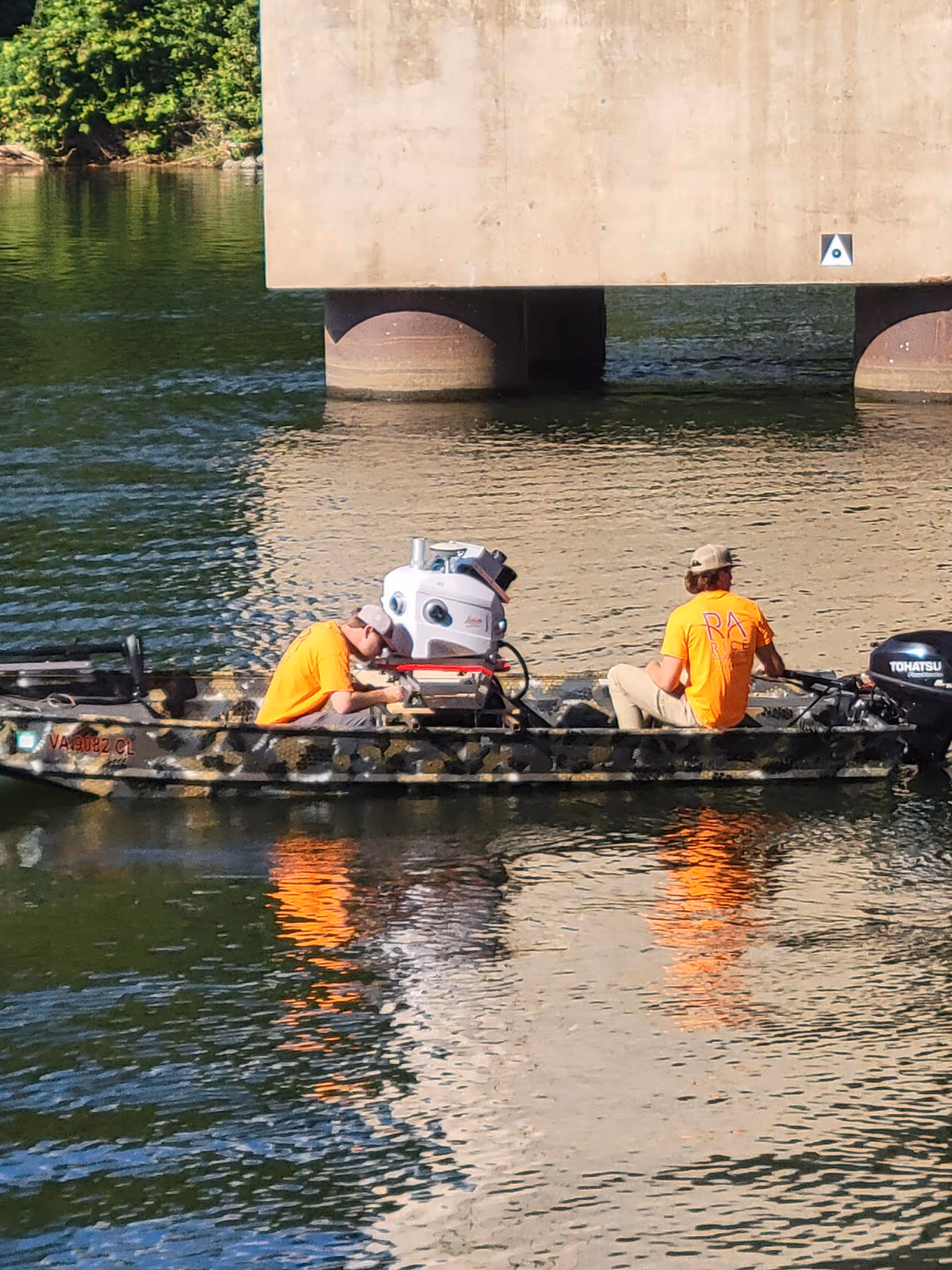 Two men in orange shirts sitting in a small boat on water near a concrete bridge support.
