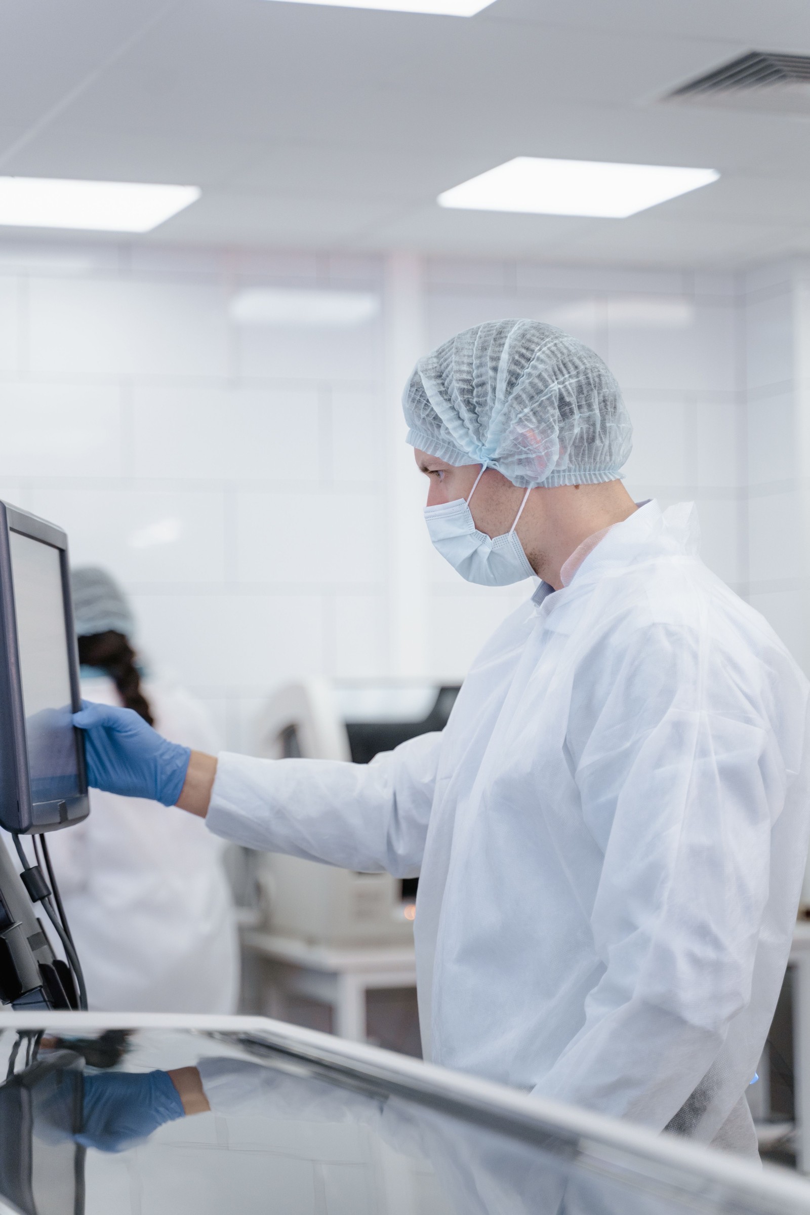 Worker wearing a hairnet, mask and gloves, operating a screen in a bright room.