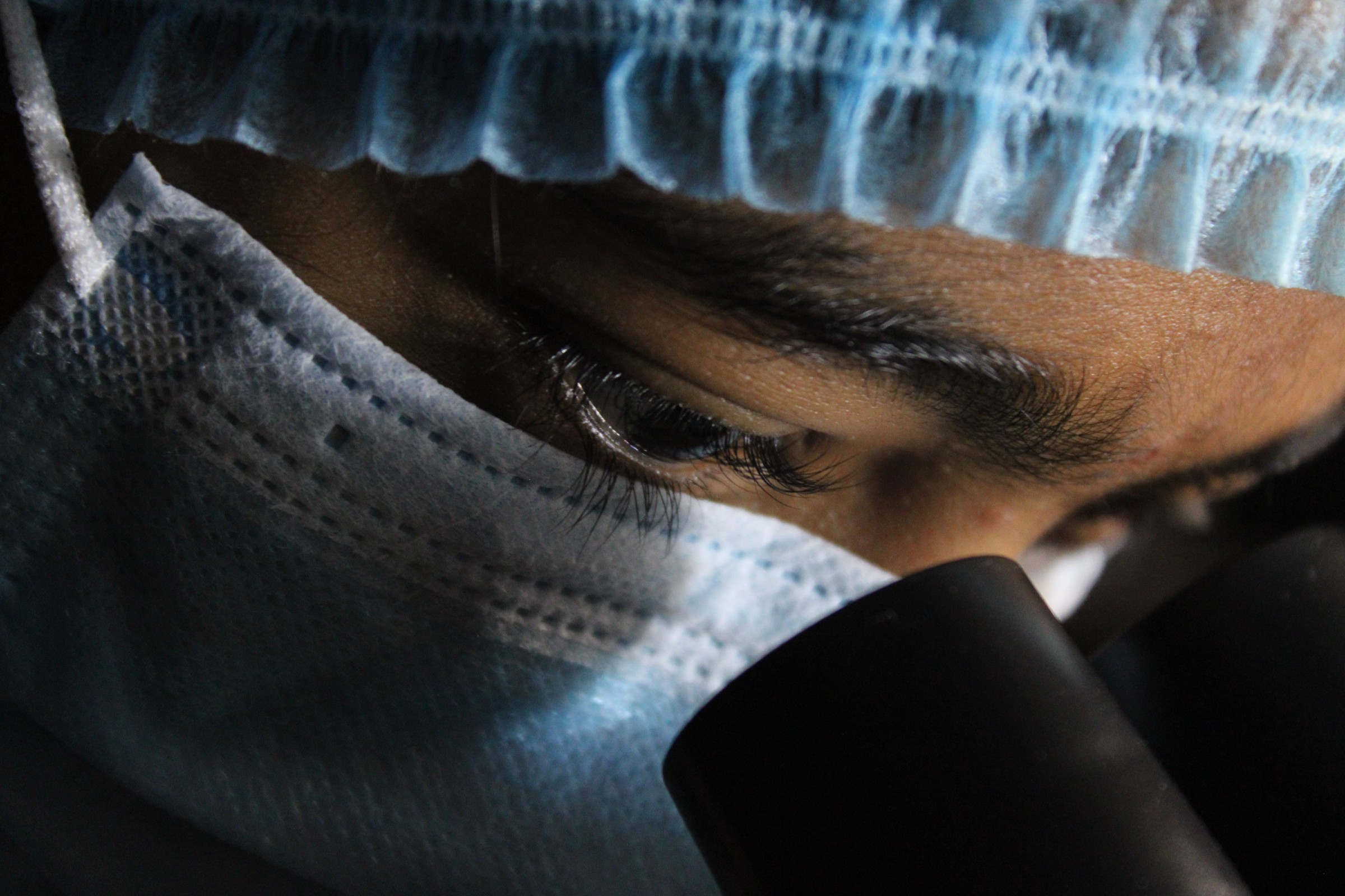 Person wearing a hairnet and mask looking through a microscope eyepiece.