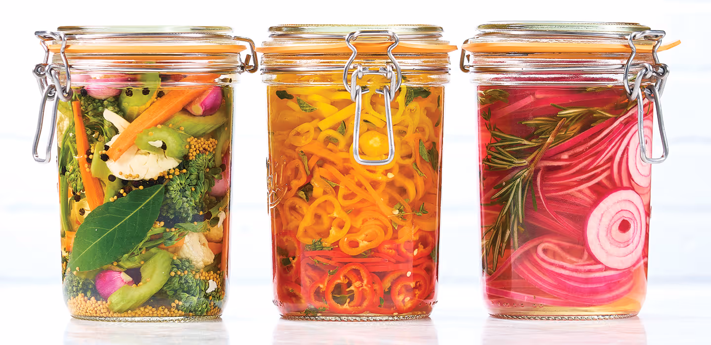 Six glass food jars full of pickled veggies and fruits including cucumbers, red onion, peppers, beets, pineapple, and broccoli.
