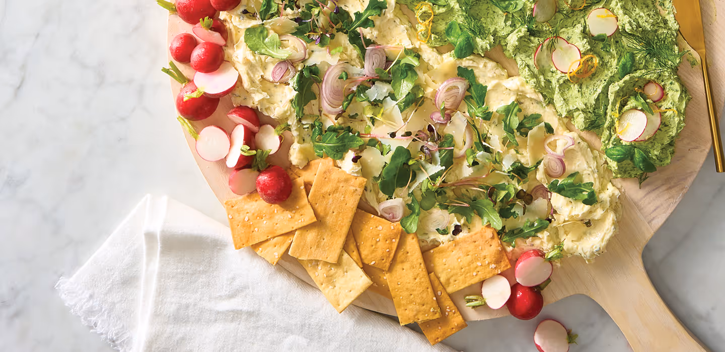 A butter board on a large round tray featuring smeared butter with fresh herbs and dippers like bread, crackers, and radishes.