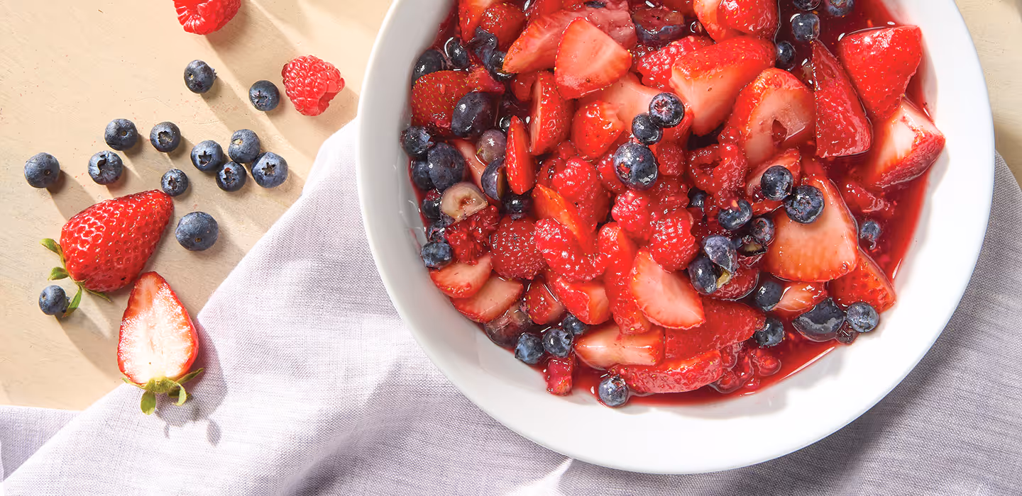 A bowl of sliced strawberries and whole raspberries and blueberries in a white bowl with a syrupy sauce.