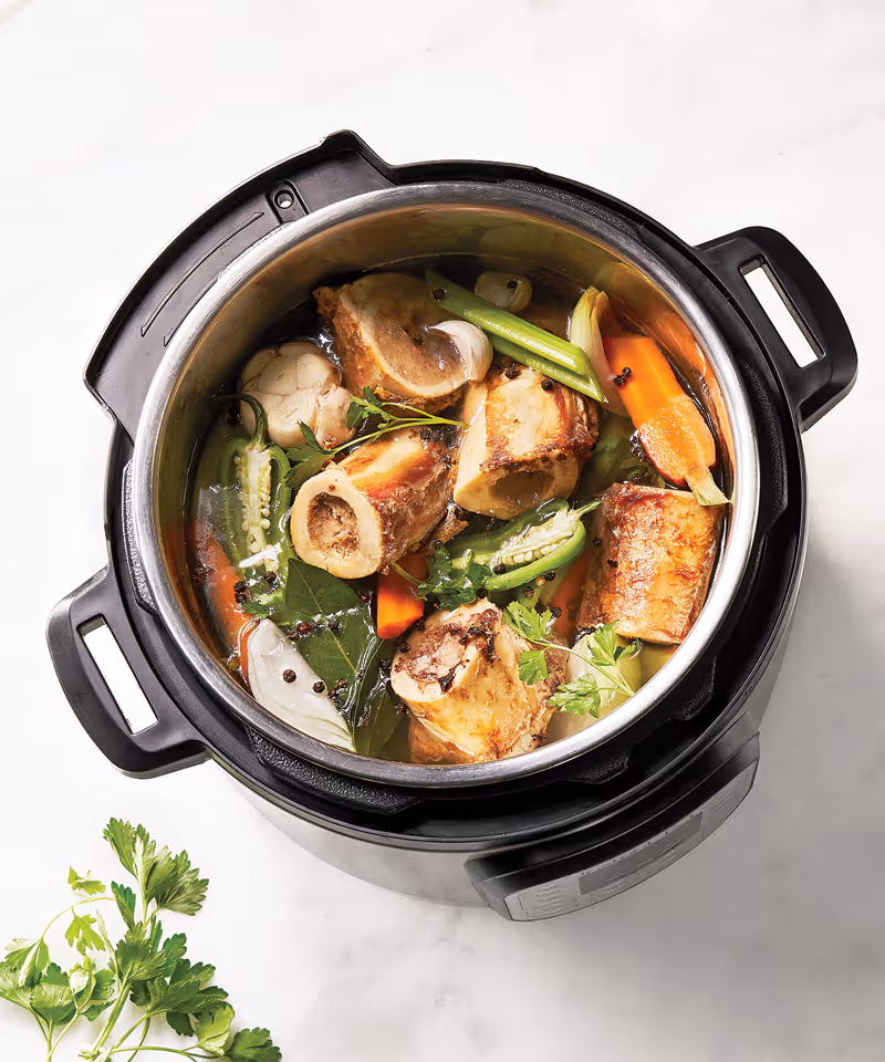 A pressure cooker full of meat, carrots, celery, jalapeño, onion, garlic, peppercorns, and fresh herbs.