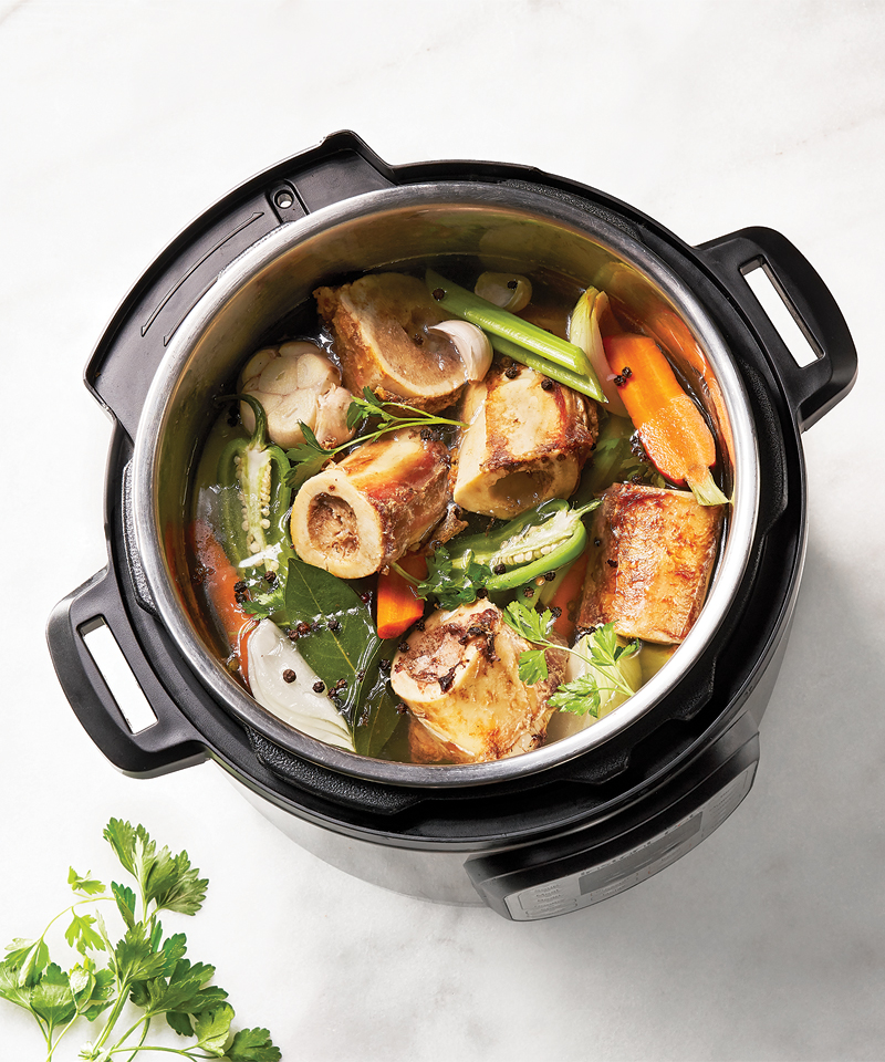 A pressure cooker full of meat, carrots, celery, jalapeño, onion, garlic, peppercorns, and fresh herbs.