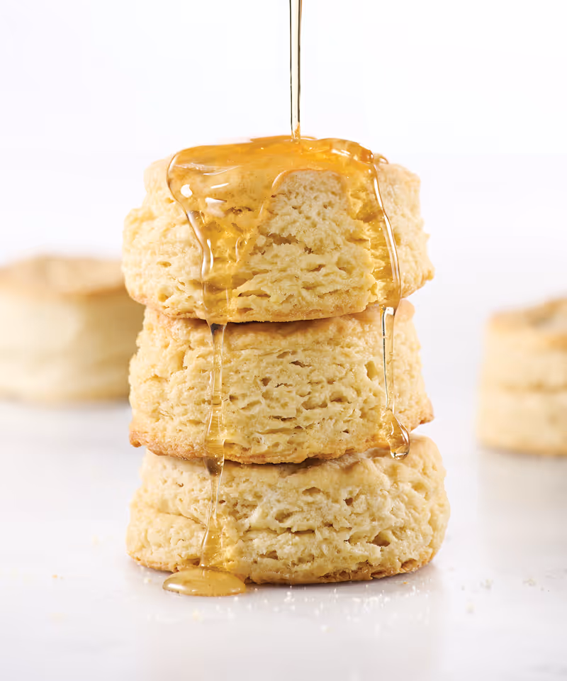 A drizzle of honey pouring onto a stack of three fluffy biscuits on a surface.