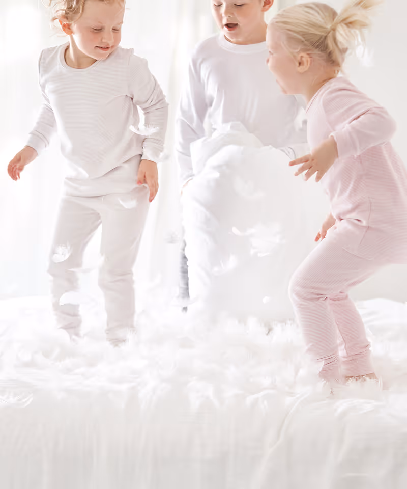 Three kids in light color clothing jumping on a bed with pillows and feathers.