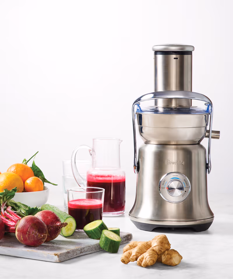 A Breville juicer with glass jars of juice and fresh whole beets, diced cucumber, oranges, and ginger on a surface.