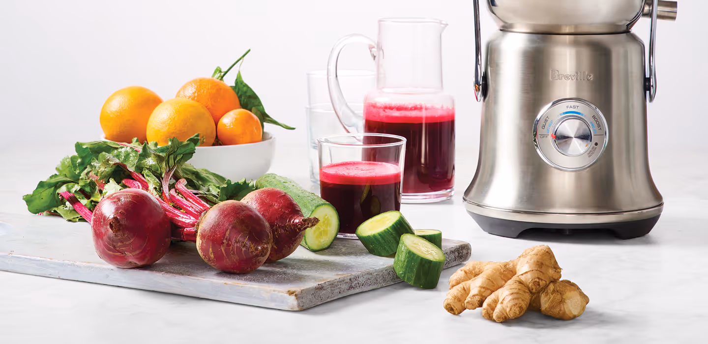 A Breville juicer with glass jars of juice and fresh whole beets, diced cucumber, oranges, and ginger on a surface.