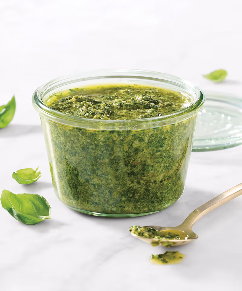 A glass jar of pesto sauce with fresh basil and a spoon on a marble surface.